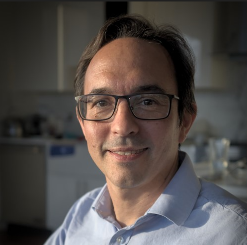 smiling man with brown side parted hair. He is clean shaven, wearing glasses and a light blue button down shirt