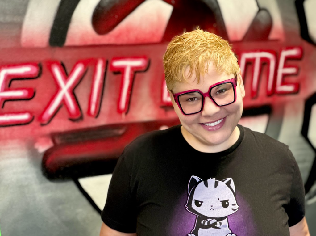 Woman with short cropped blonde hair, smiling, wearing large red and black plastic frame glasses. She is wearing a black t-shirt with a grumpy cartoon cat and standing in front of a wall that says "exit game"