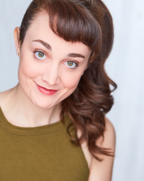 smiling woman with hazel eyes and chestnut hair in a curled ponytail and bangs. She is wearing an olive green sleeveless shirt