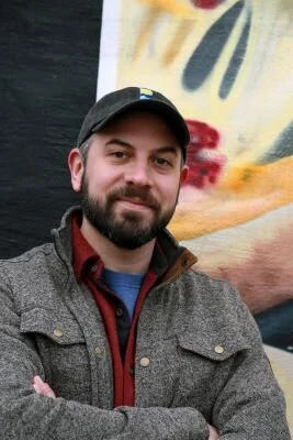 man with thick dark beard, wearing black baseball cap and wool shirt, smiling.